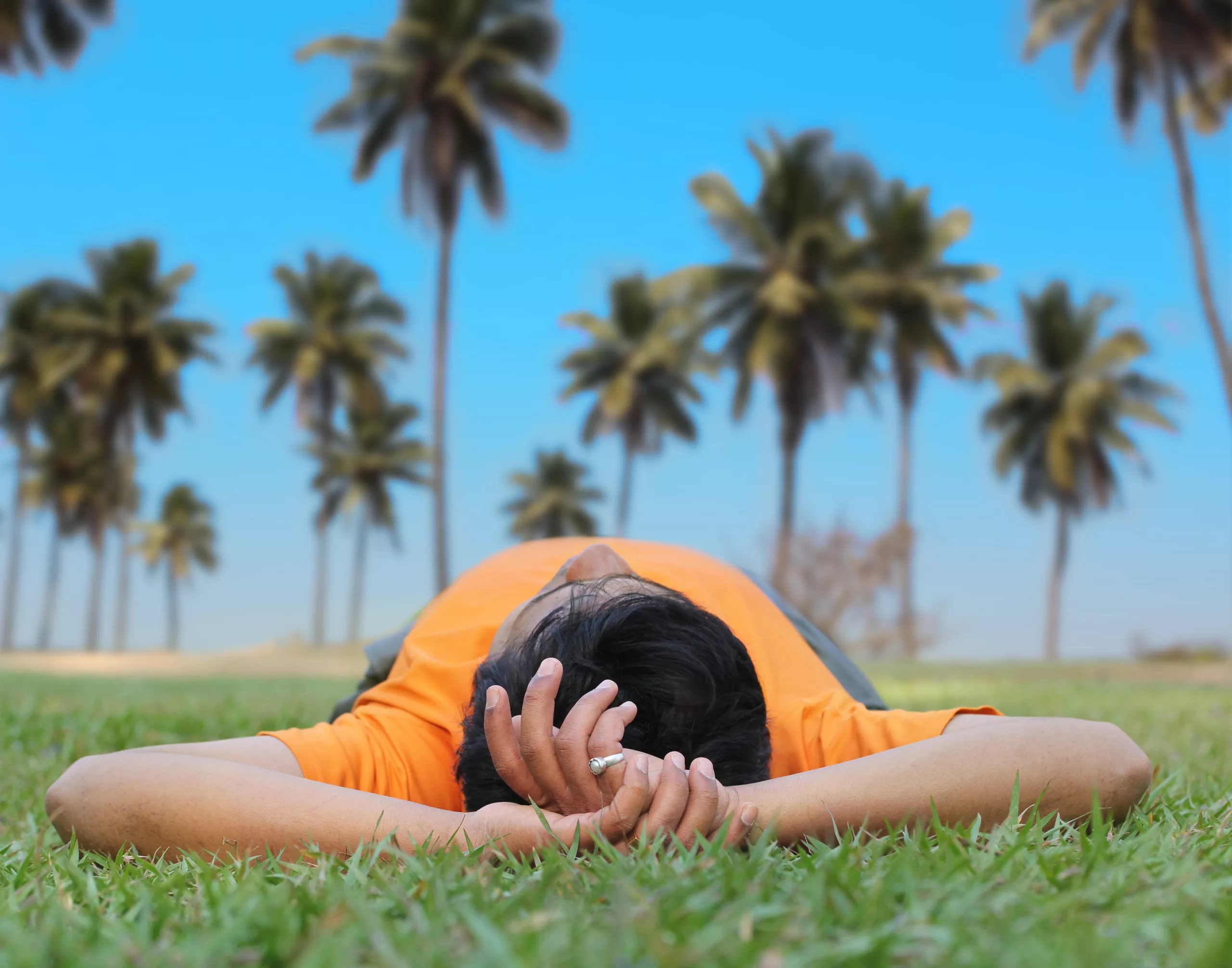 A young man relaxes his mind from stress while lying on the grass, with his hands behind his head, staring up at a bright blue sky and palm trees