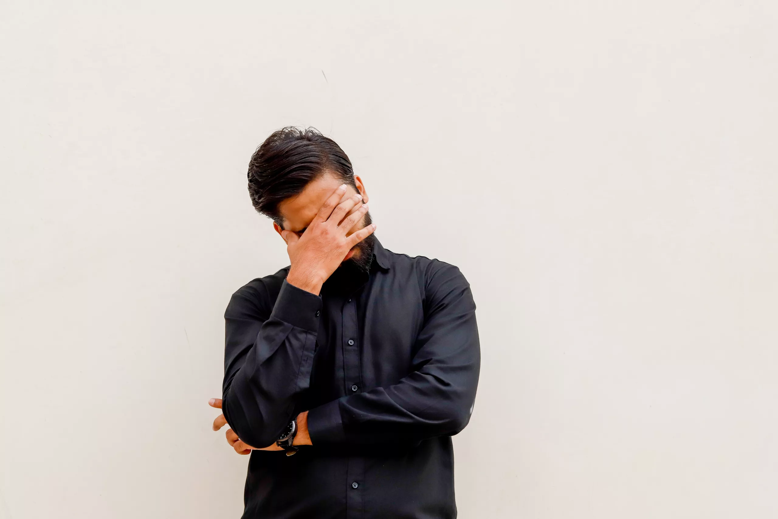 A south asian man in a black shirt stands against a gray background. He hugs himself with one arm, and with the other, covers his face.
