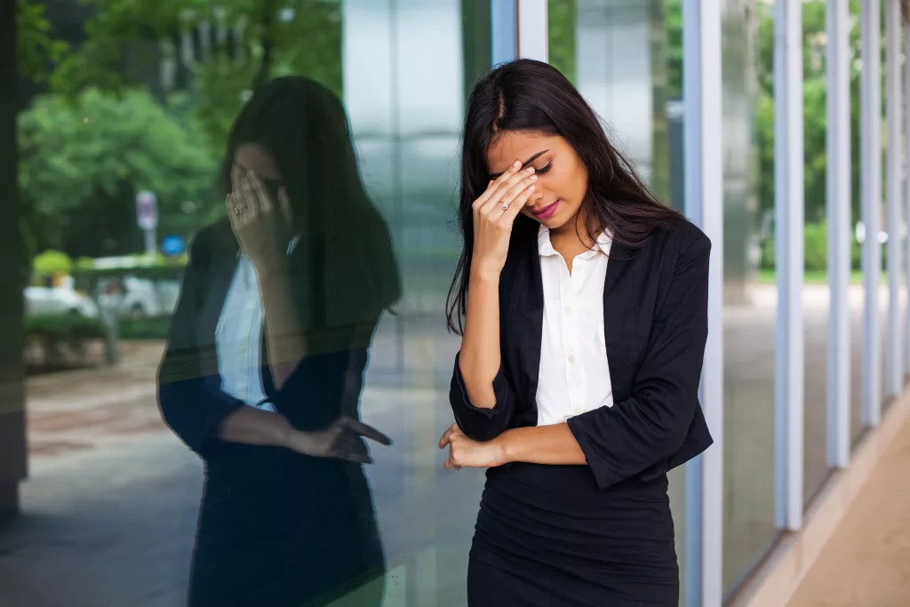 A stressed indian business woman leans against the glass windows of an office building, holding her head as if it aches