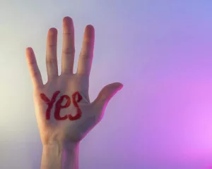 a hand raised against a blurred purple background, with 'yes' written on the palm in red ink